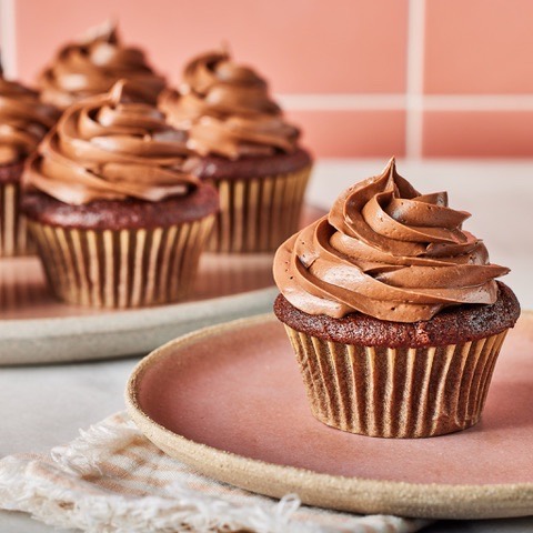 A close-up of a delicious chocolate cupcake with a swirl of frosting on a pink plate, with several cupcakes in the background.