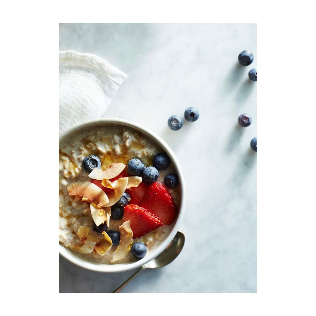 Overhead shot of oatmeal bowl with sliced strawberries, blueberries, coconut flakes, and syrup drizzle on marble surface.