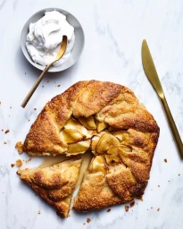 Top-down view of a golden baked apple galette with a slice cut out, alongside a bowl of whipped cream and a brass knife on a marble surface.