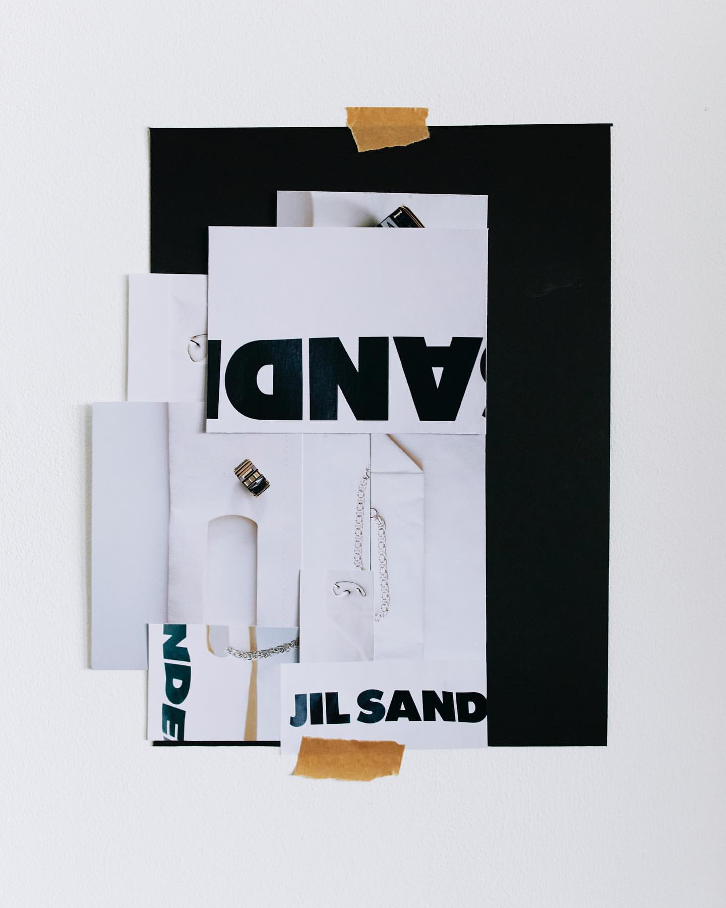 Abstract collage on white backdrop featuring a black rectangle, white paper scraps, Jil Sander logo, and rings adhered with tape.