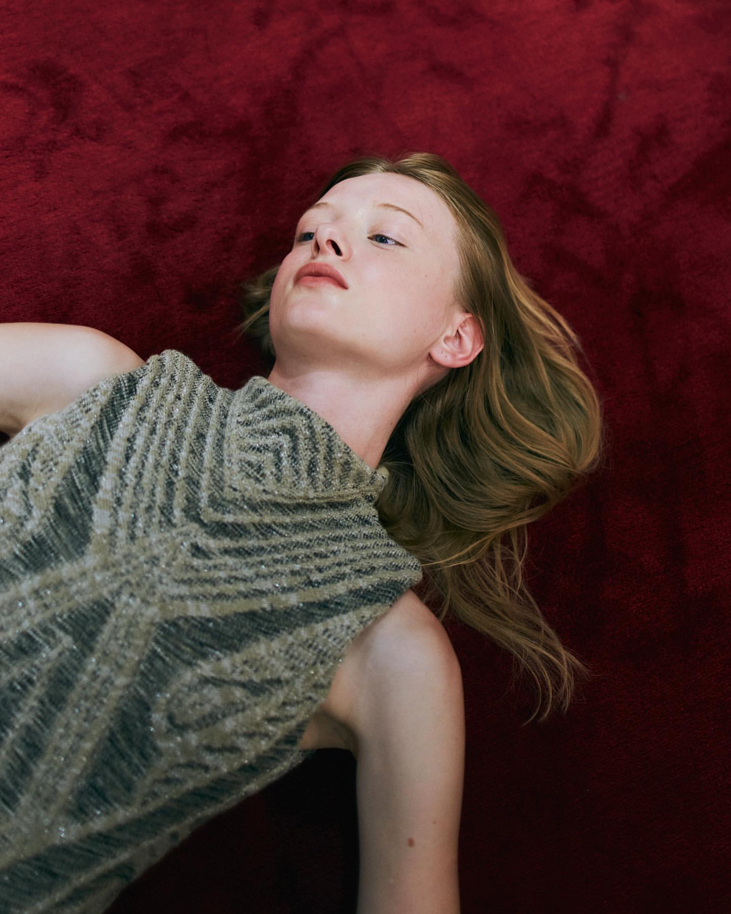 Portrait of a fair-skinned young adult woman lying on a red textured surface, wearing a gray knitted top with geometric patterns.