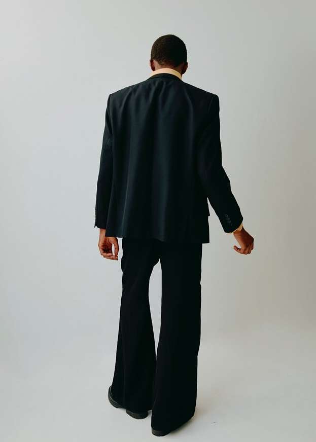 A man from the back, dressed in a suit and standing against a white background in a studio. He is wearing flared pants.