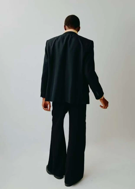 A man from the back, dressed in a suit and standing against a white background in a studio. He is wearing flared pants.