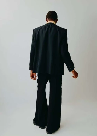 A man from the back, dressed in a suit and standing against a white background in a studio. He is wearing flared pants.