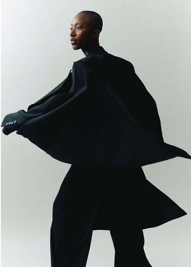 Elegant African woman in flowing black coat and wide-leg pants, posing against white backdrop. Studio lighting accentuates her sleek, shaved hair.