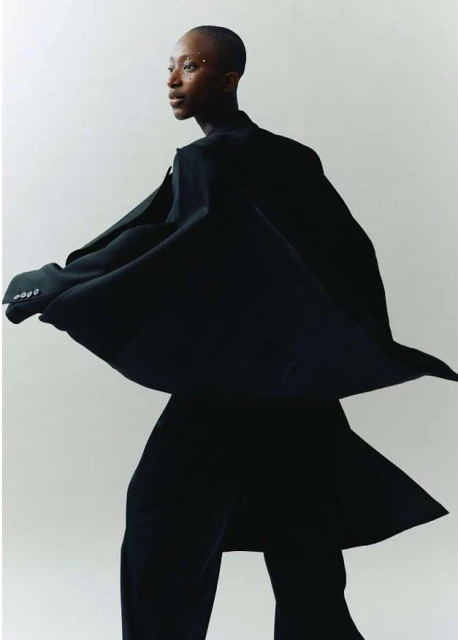 Elegant African woman in flowing black coat and wide-leg pants, posing against white backdrop. Studio lighting accentuates her sleek, shaved hair.