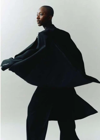 Elegant African woman in flowing black coat and wide-leg pants, posing against white backdrop. Studio lighting accentuates her sleek, shaved hair.