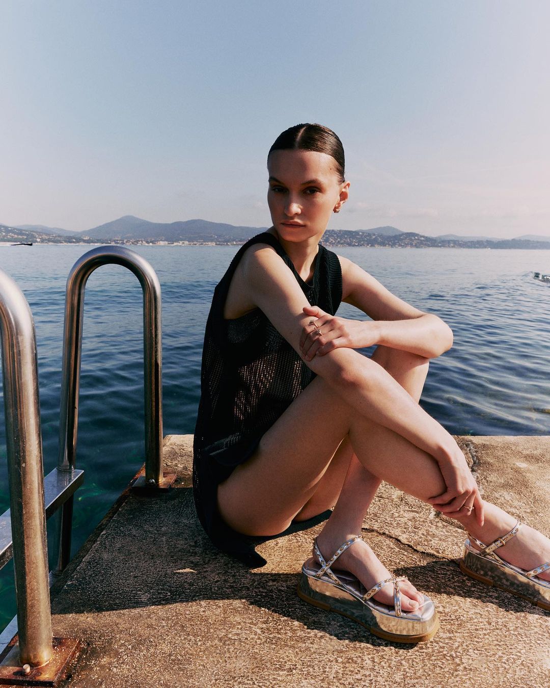 Woman in black top and platform sandals poses on dock with ocean and mountains in background on sunny day, fashion shoot.