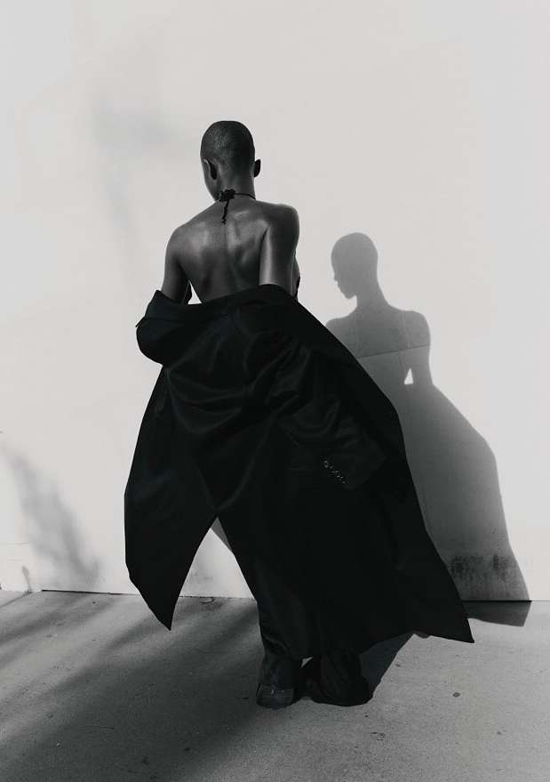 Woman in fashionable black coat poses against a white wall with her shadow, a black-and-white studio portrait.
