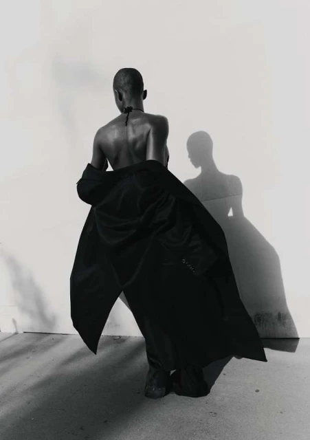 Woman in fashionable black coat poses against a white wall with her shadow, a black-and-white studio portrait.