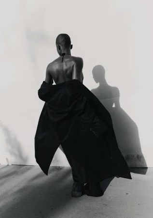 Woman in fashionable black coat poses against a white wall with her shadow, a black-and-white studio portrait.