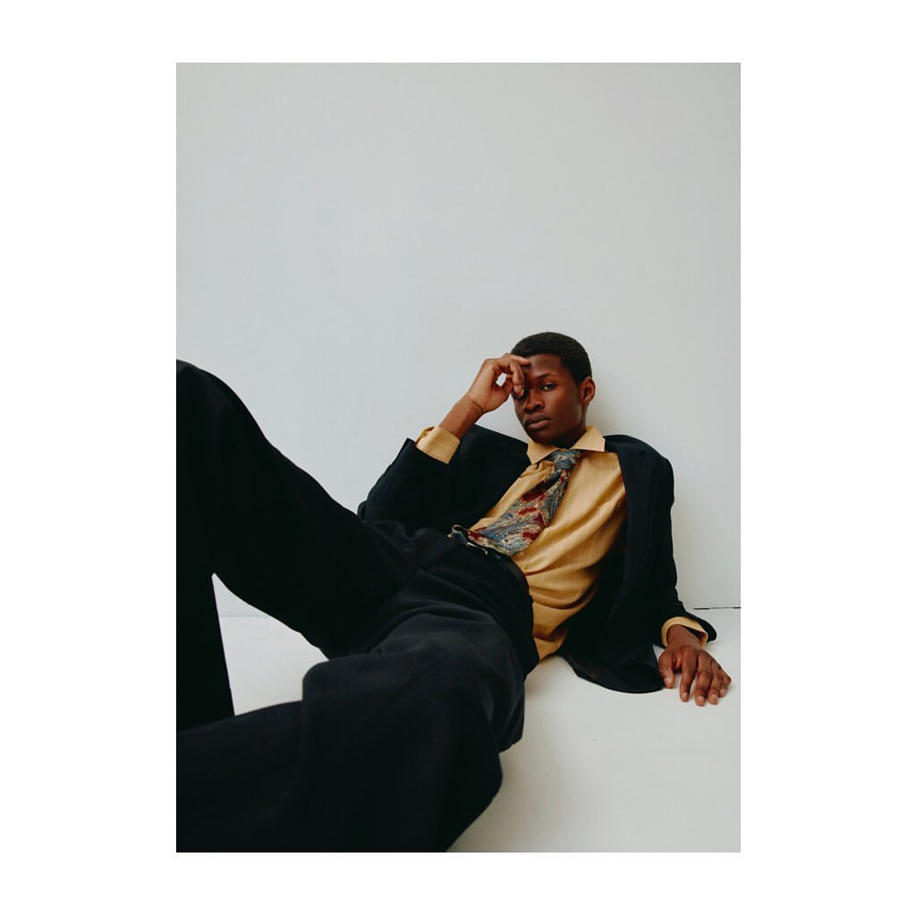 Dark-skinned male model in a blazer, shirt, and tie reclines against a white backdrop in a fashion-style portrait. Neutral color grading.
