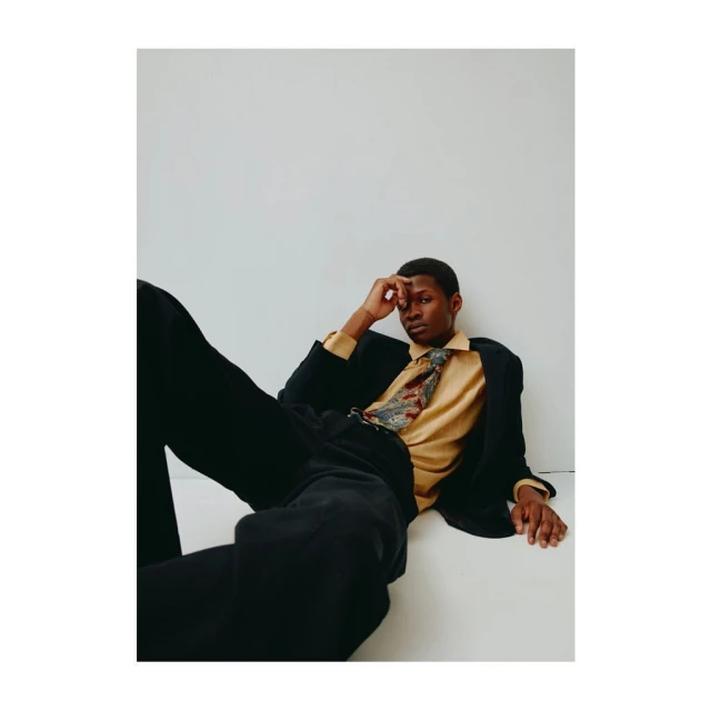 Dark-skinned male model in a blazer, shirt, and tie reclines against a white backdrop in a fashion-style portrait. Neutral color grading.