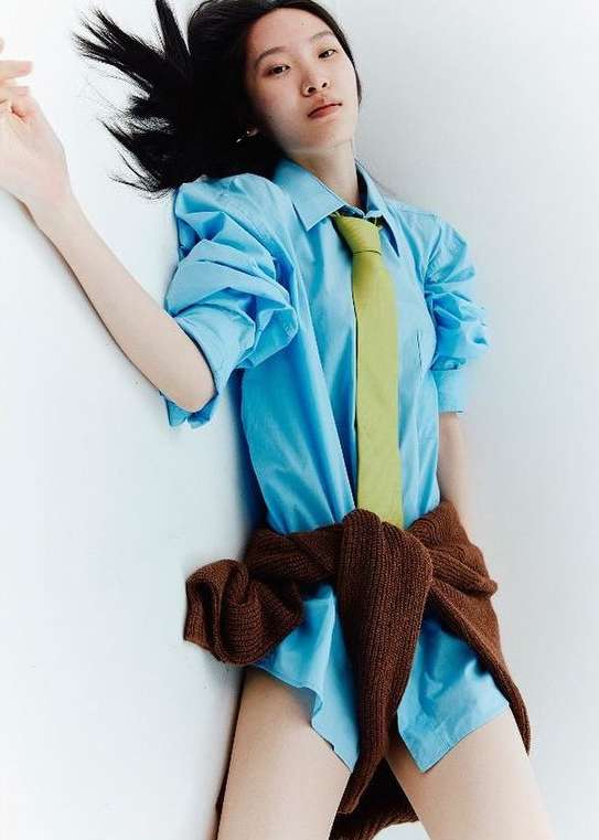Woman with black hair poses lying against a white surface wearing a blue button-down shirt and green tie with a brown sweater tied around her waist.