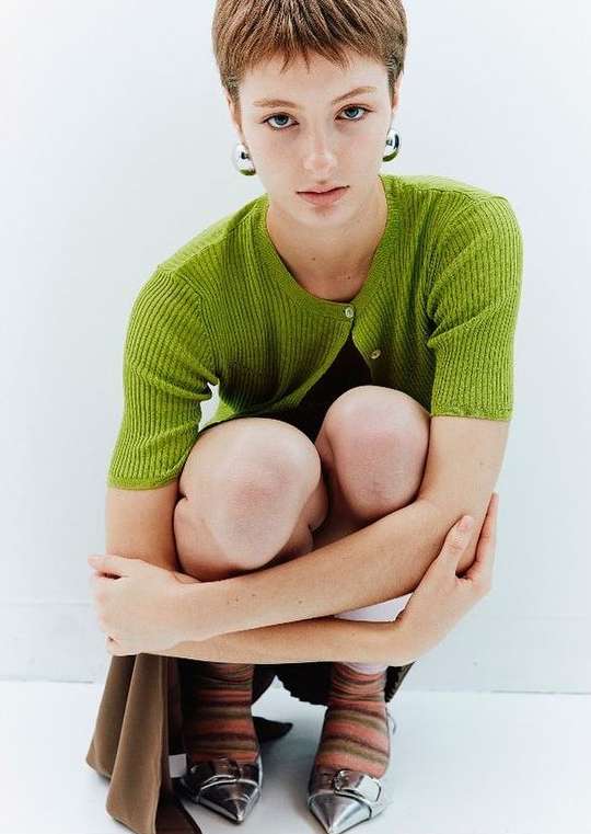 A young woman with short brown hair poses in a green ribbed shirt, brown skirt, striped socks, and pointy silver shoes against white backdrop.