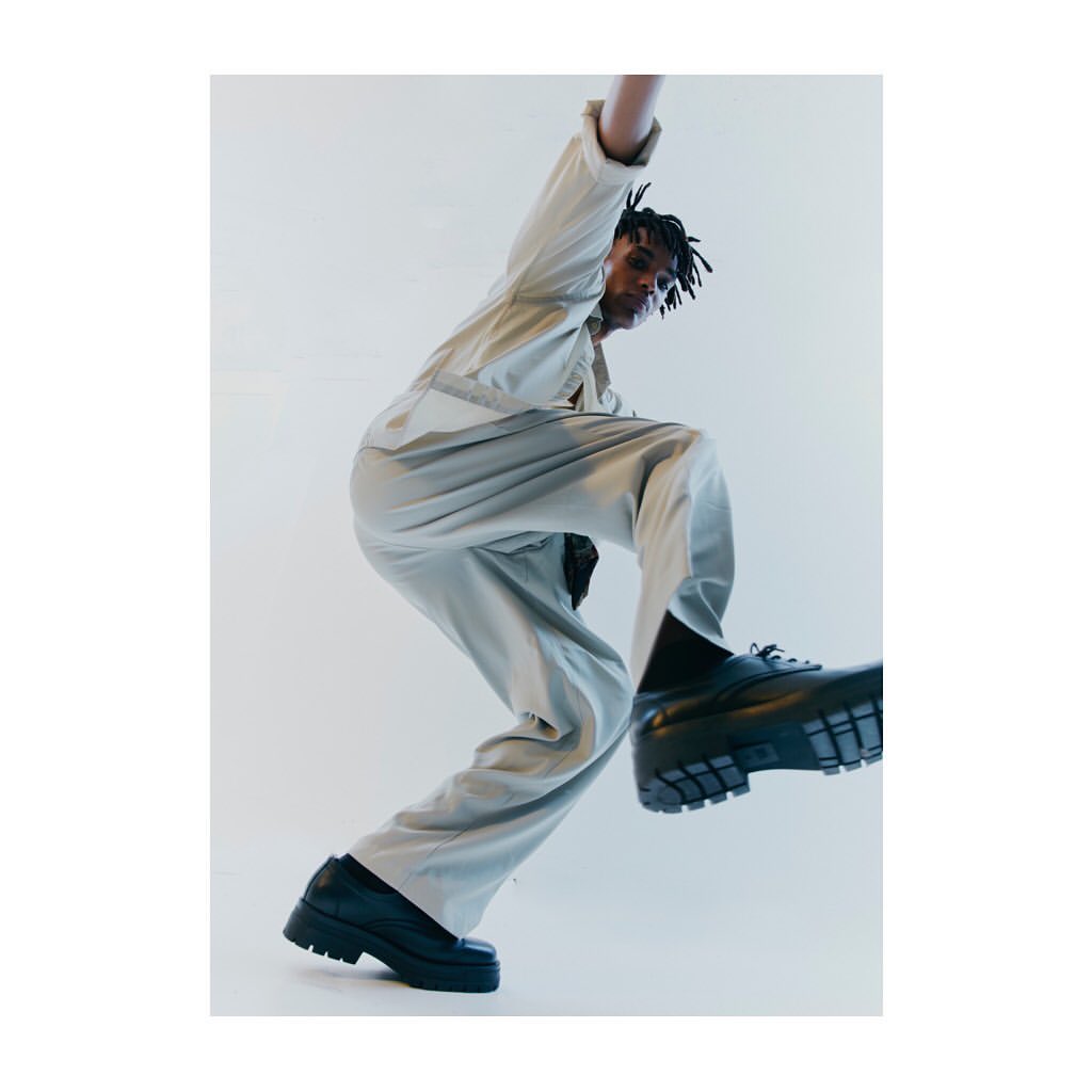 Low-angle shot of an African American man in a beige jumpsuit and black platform shoes leaping against a white backdrop in a photo studio.