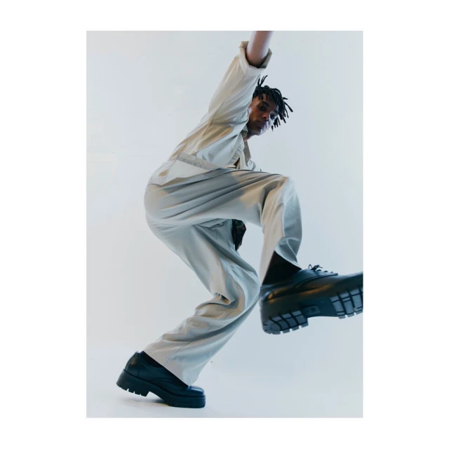 Low-angle shot of an African American man in a beige jumpsuit and black platform shoes leaping against a white backdrop in a photo studio.