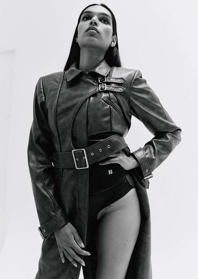 Monochrome fashion shot of a slender model wearing a belted leather trench coat and black bodysuit against a bright, minimalist background.