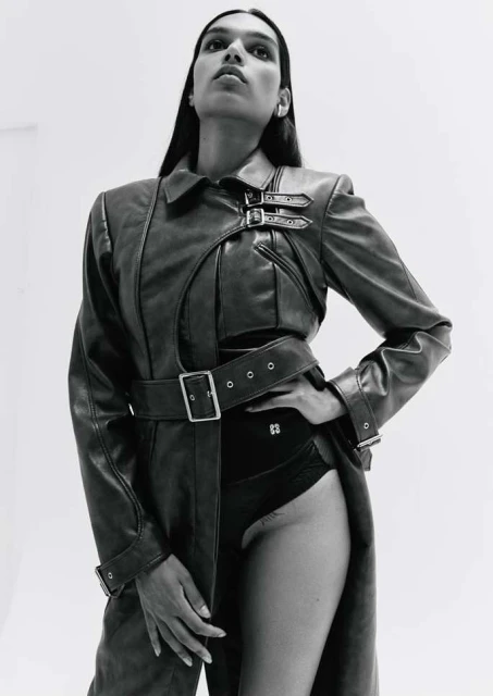 Monochrome fashion shot of a slender model wearing a belted leather trench coat and black bodysuit against a bright, minimalist background.