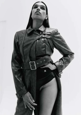 Monochrome fashion shot of a slender model wearing a belted leather trench coat and black bodysuit against a bright, minimalist background.