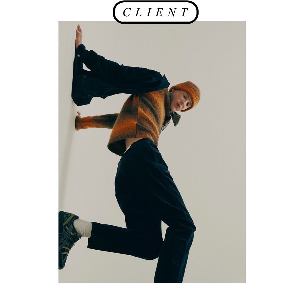 A young adult poses in an orange and brown sweater and blue pants against a plain background in an eye-level studio shot.
