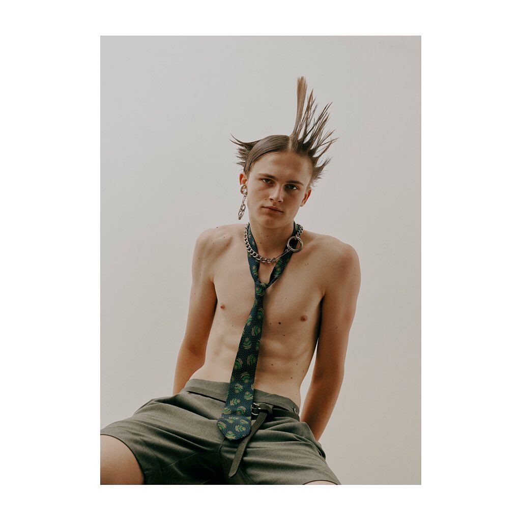 A shirtless young adult male with a mohawk hairstyle and a tie poses in a studio with a neutral background.
