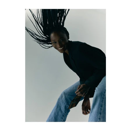 Smiling Black woman with long braided hair whipping through the air wearing a dark blazer and blue jeans
