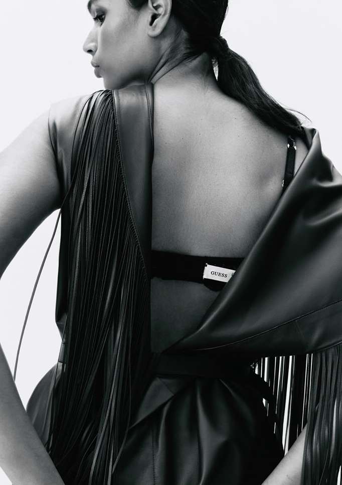 Woman in leather fringe dress shot from behind in a studio. Dark hair pulled back and styled, looking off-camera, soft lighting.