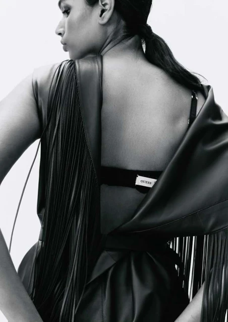 Woman in leather fringe dress shot from behind in a studio. Dark hair pulled back and styled, looking off-camera, soft lighting.