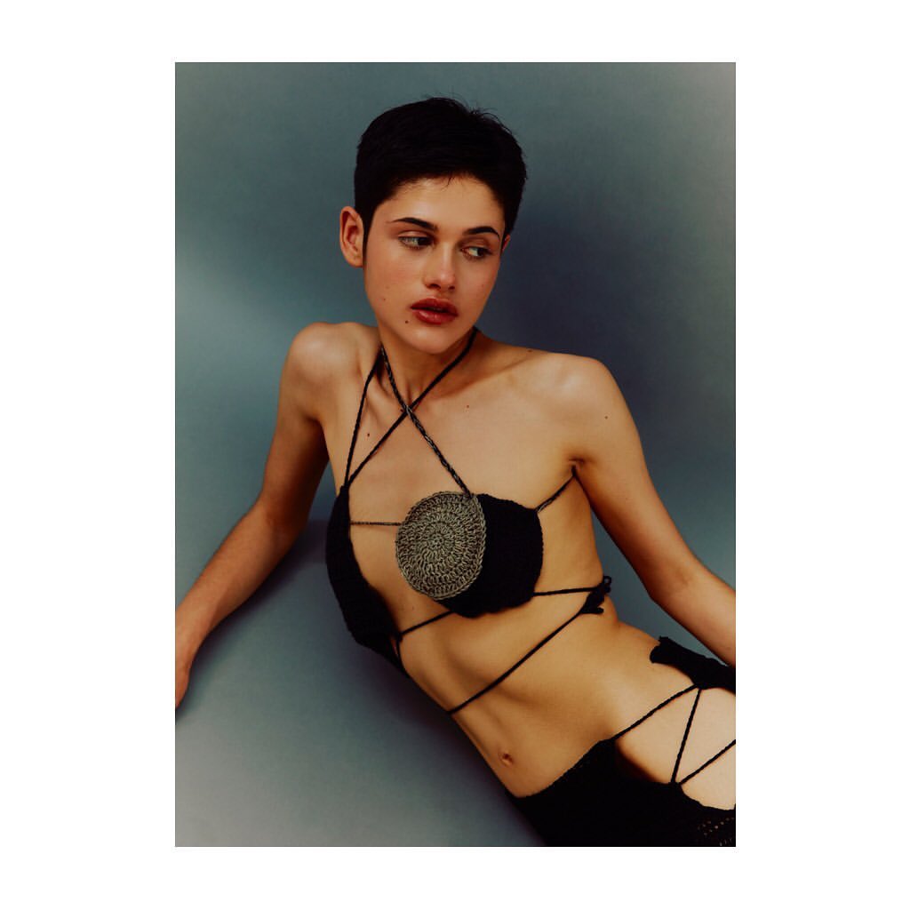 Portrait of a young woman with short dark hair wearing a black crocheted outfit against a solid gray background in a fashion editorial.