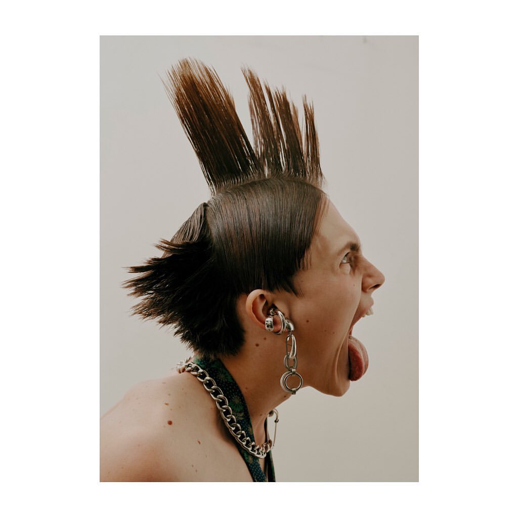 Profile portrait of a young adult with a mohawk hairstyle, open mouth, and exposed tongue, wearing jewelry against an off-white background.