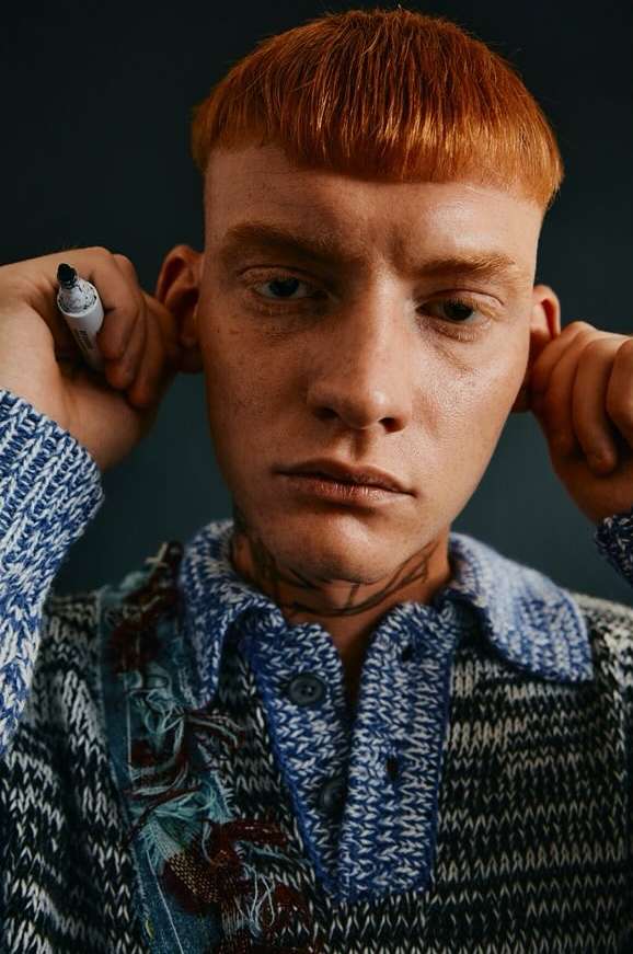 Close-up shot of a fair-skinned adult male with short reddish hair, holding a marker, wearing a patterned blue and gray sweater.