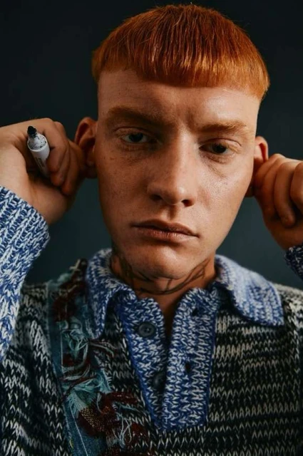 Close-up shot of a fair-skinned adult male with short reddish hair, holding a marker, wearing a patterned blue and gray sweater.