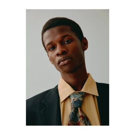 Portrait of a Black man in a dark suit jacket, yellow striped shirt, and patterned tie against a light grey background.