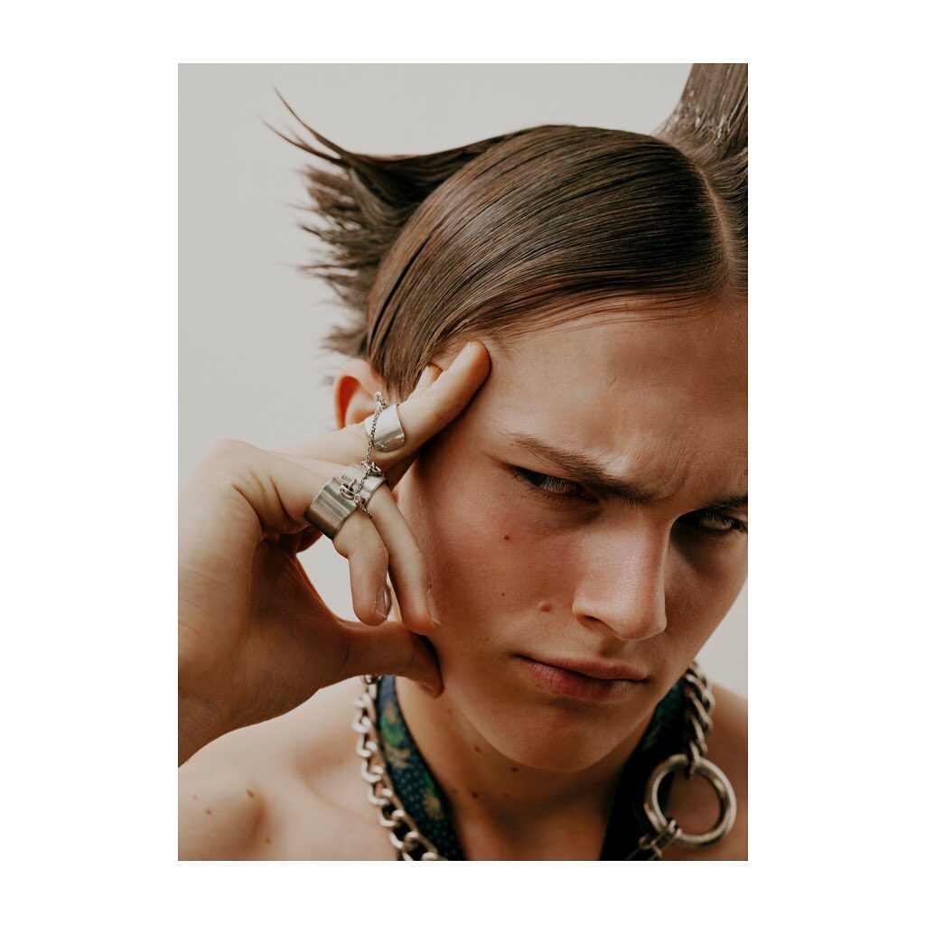 Dramatic close-up of a young adult male with slicked-back, spiky hair, wearing silver rings and a chain necklace against a light background.
