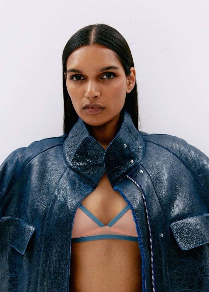 Close-up studio shot of a dark-skinned woman wearing a blue leather jacket and pink bra, posing against a white background.
