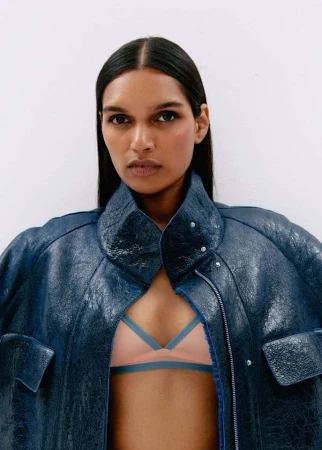Close-up studio shot of a dark-skinned woman wearing a blue leather jacket and pink bra, posing against a white background.