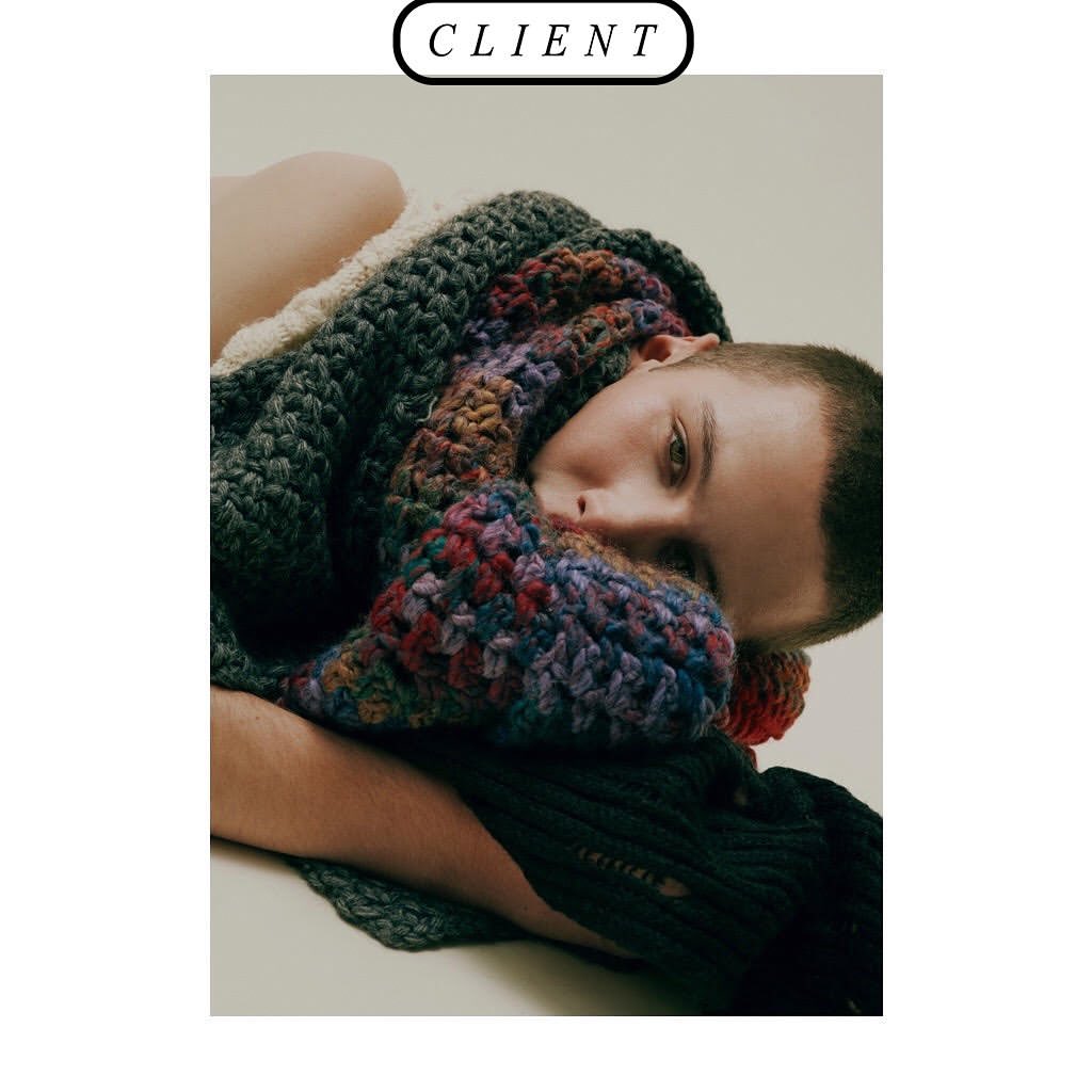Androgynous person posing while wrapped in a multi-colored, chunky knit scarf indoors against a neutral studio backdrop with natural light.