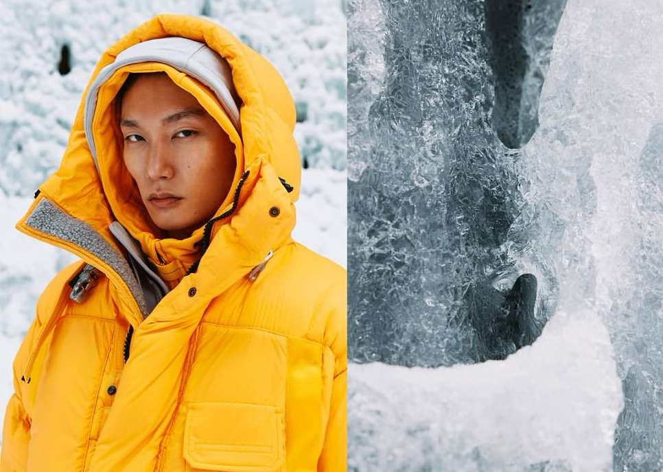 Asian male model in a bright yellow puffer jacket poses in front of a blue glacier with textured ice for a fashion editorial shoot.