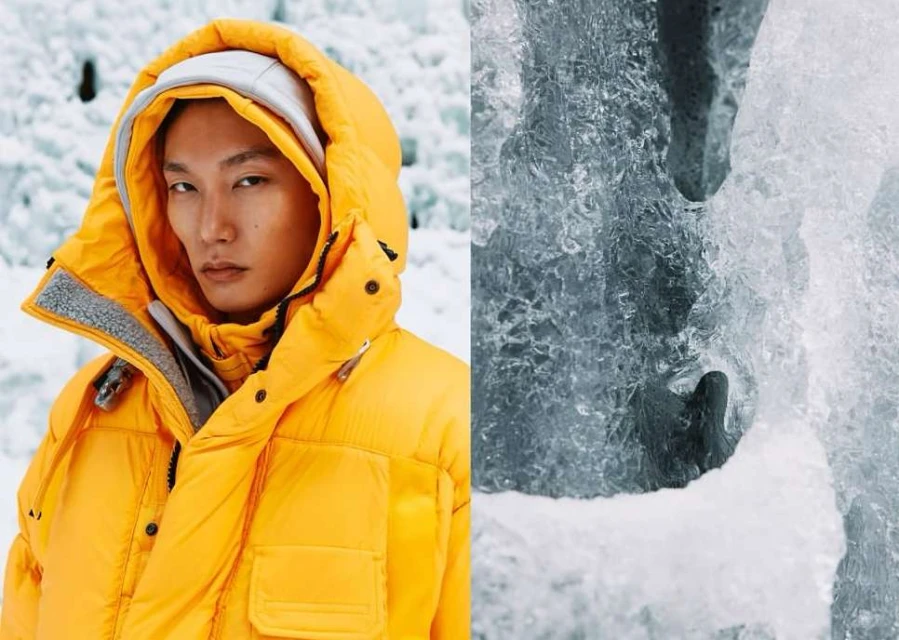 Asian male model in a bright yellow puffer jacket poses in front of a blue glacier with textured ice for a fashion editorial shoot.
