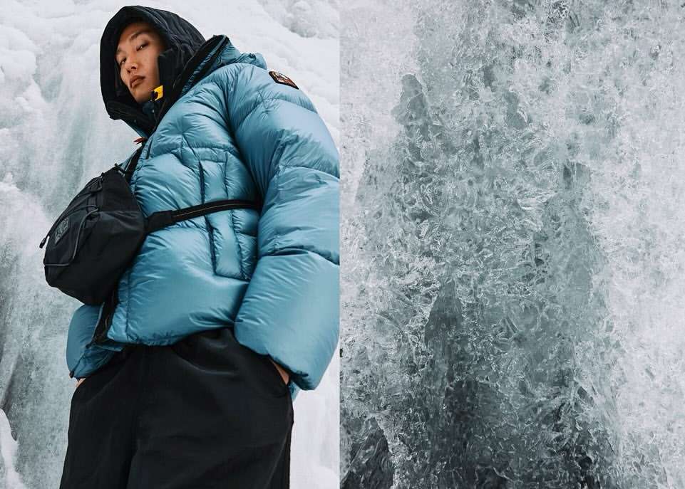 Asian man in a blue puffer jacket and black bag stands against an icy waterfall. Winter fashion and nature merge in this shot.