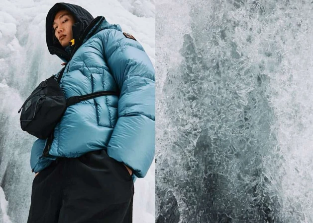 Asian man in a blue puffer jacket and black bag stands against an icy waterfall. Winter fashion and nature merge in this shot.