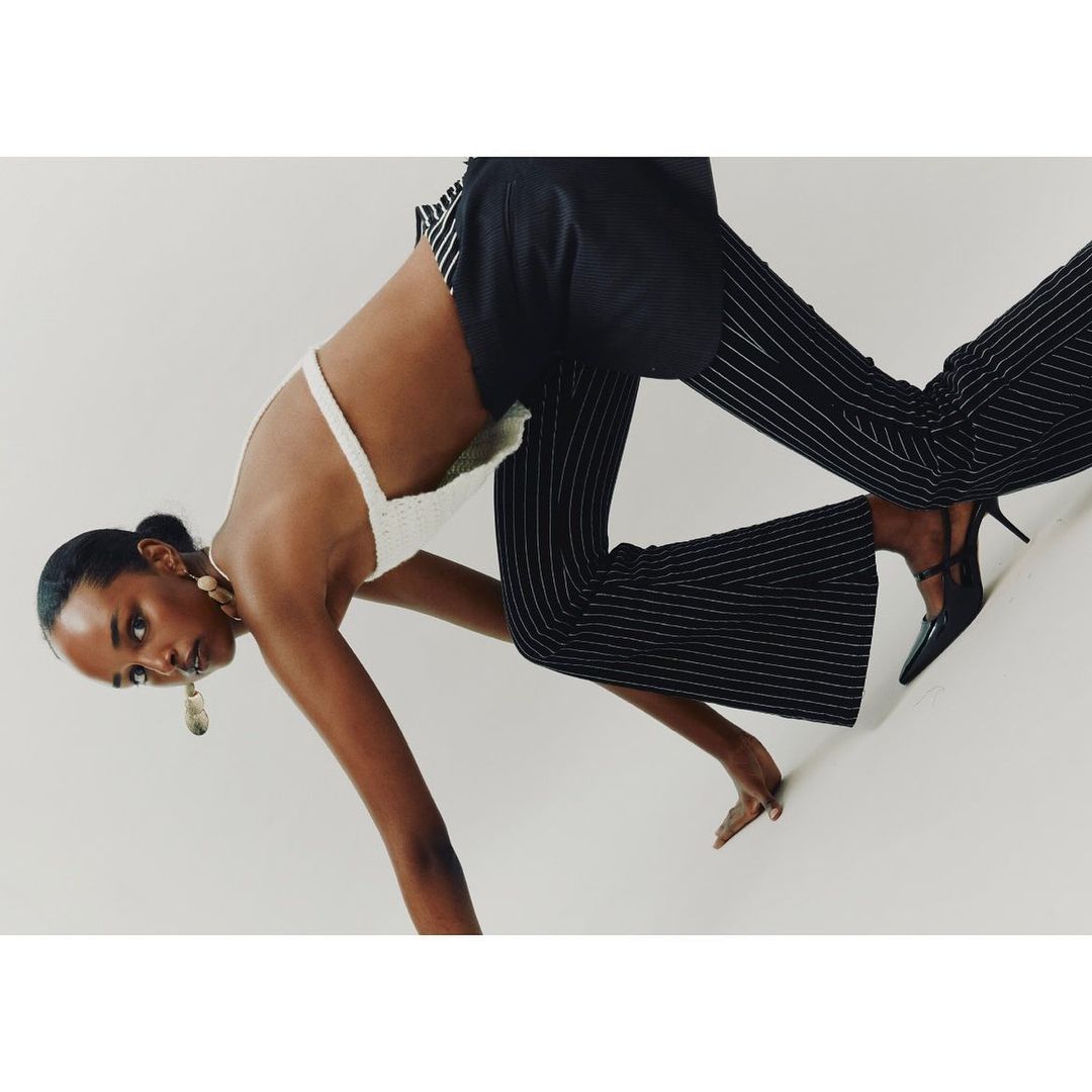 Fashion shot on an angle featuring a woman in a white backless top, dark striped pants, and high heels against a stark white backdrop.