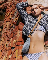 Model wearing a blue patterned jacket and bikini posing against a textured red brick and tile wall outdoors in bright sunlight