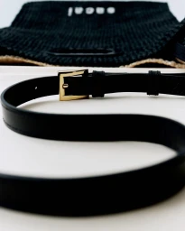 Close-up of a black leather belt with a gold buckle on a white surface with a woven bag in the background