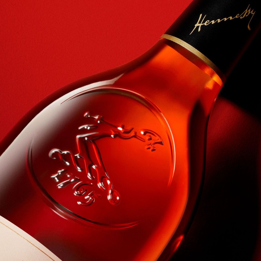 Close-up shot of a Hennessy bottle with the logo embossed on the glass, against a vibrant red backdrop, highlighting the brand's elegance.
