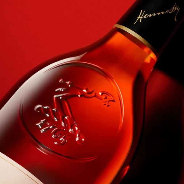 Close-up shot of a Hennessy bottle with the logo embossed on the glass, against a vibrant red backdrop, highlighting the brand's elegance.