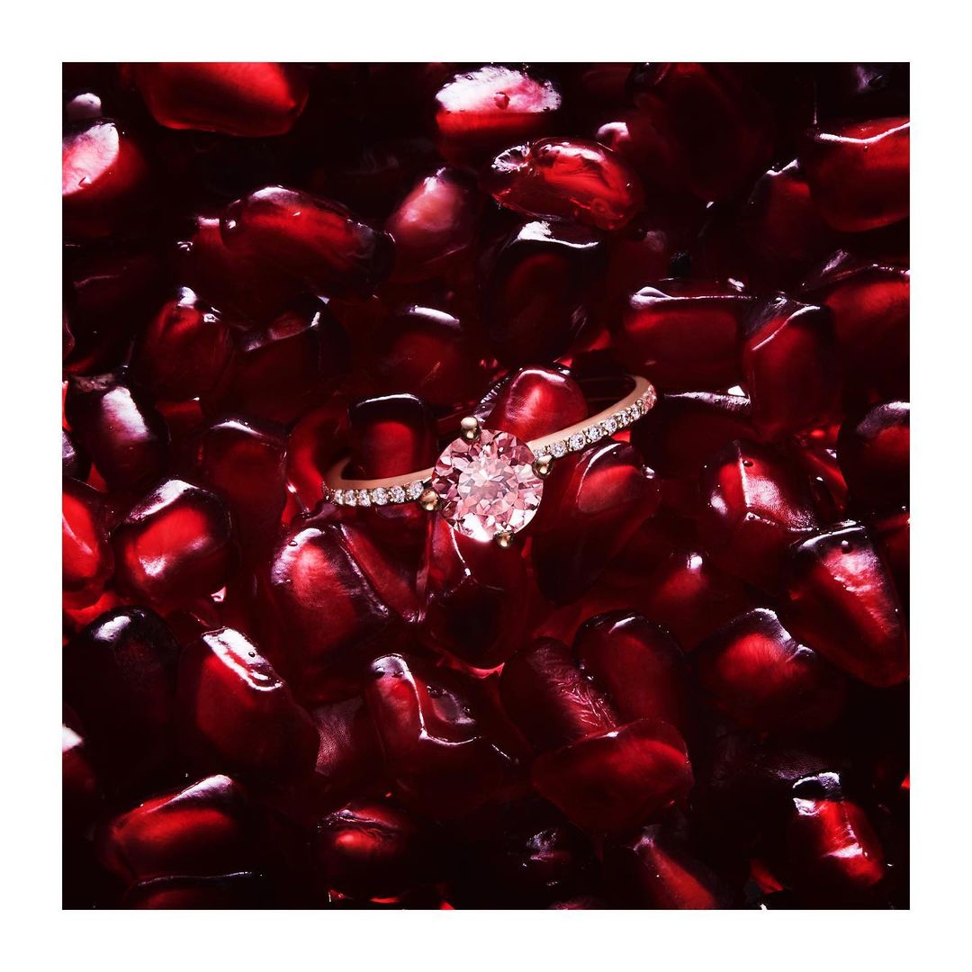 Engagement ring on a bed of vibrant pomegranate seeds. The ring features a round stone and sits atop the deep red seeds.