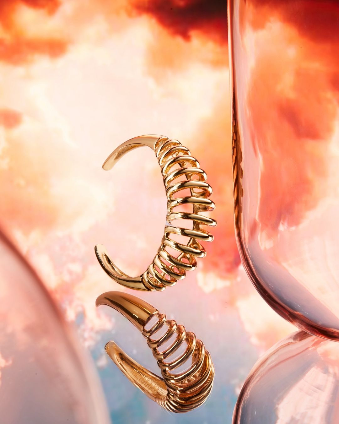 Elevated shot of a gold cuff bracelet reflecting against orange, pink, and white cloudscape, styled with a tall glass vase nearby.