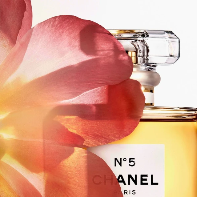 Close-up of a perfume bottle, partially obscured by a pink flower petal, with visible Chanel No. 5 logo against a white background.